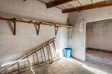 An old, unfinished room with white walls, a wooden shelf, a ladder leaning, a blue bucket, and exposed ceiling beams. Sunlight hits the floor.