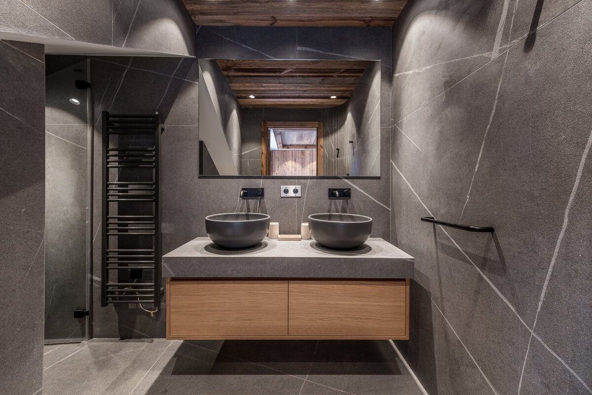 Modern bathroom with dark grey stone walls, a floating wood vanity, two bowl sinks, and a large rectangular mirror.