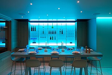 A modern bar and dining area with striking blue lighting. Illuminated shelves hold bottles/glasses, and a long table is set for meals with multiple chairs.