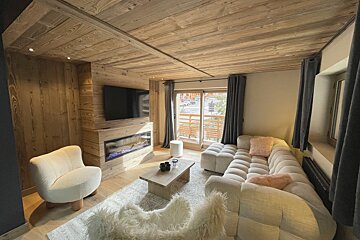 A rustic-chic living room features wood paneling, a white sectional sofa, TV above an electric fireplace, and a balcony view.