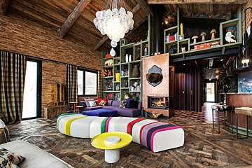 A vibrant, eclectic chalet living room with rustic wood features, a unique balloon chandelier, colorful seating, a modern fireplace, and tall bookshelves.