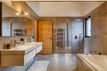 A sleek, modern bathroom featuring double sinks, a glass shower, bathtub, and a warm wooden door, complemented by gray tiled walls and floor.