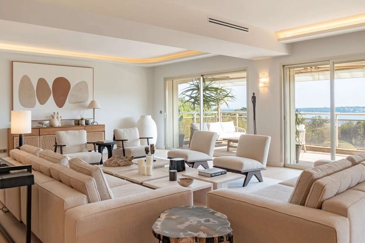 A spacious, luxurious living room in neutral tones with large sofas, artistic decor, and a balcony offering a stunning view of the sea or lake.