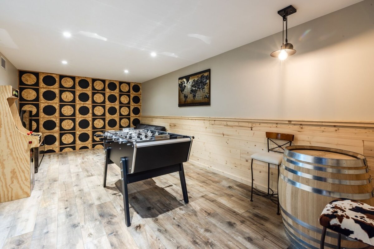 A stylish game room featuring a foosball table, an arcade cabinet, wood-paneled walls, and a barrel table with bar stools.