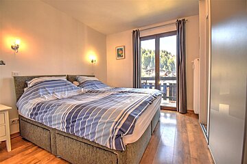 A warm bedroom features a large bed with blue striped bedding, wooden floors, wall sconces, and a balcony overlooking a lush mountain view.