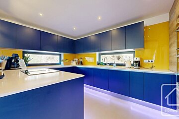 Sleek blue and yellow modern kitchen with white countertops, ambient under-cabinet lighting, and horizontal windows overlooking greenery.