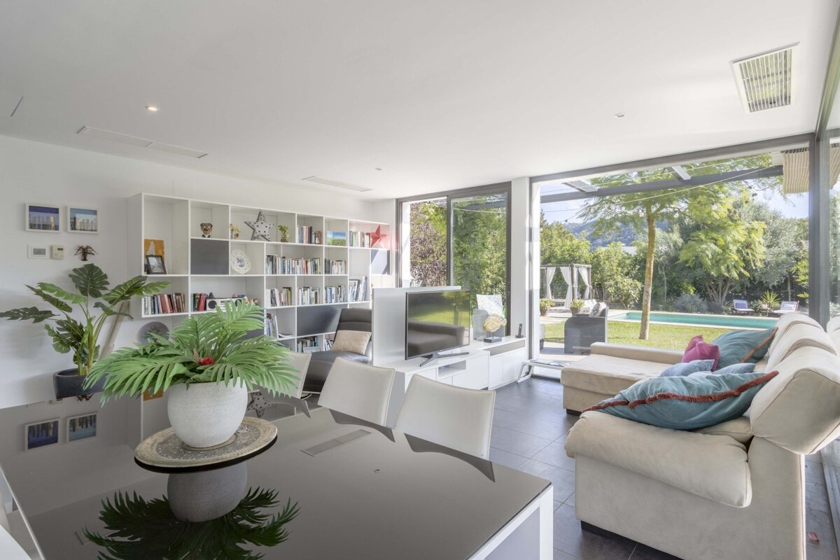 A bright, modern living/dining room features white bookshelves, TV, and comfortable seating, with large glass doors opening to a lush garden and pool.