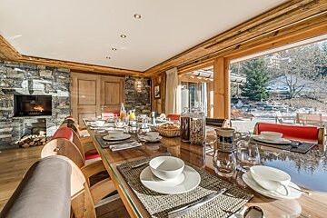 A cozy dining room features a long table set for breakfast, a crackling fireplace, and large windows revealing a snowy outdoor landscape.