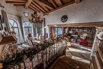 Luxurious chalet interior featuring a grand dining area, cozy lounge with a bar, vaulted wooden ceiling, and a festive garland on the railing.