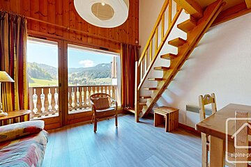 Chalet-style room with wood paneling, a wooden staircase, and a balcony offering stunning mountain valley views. Features simple wooden furniture.