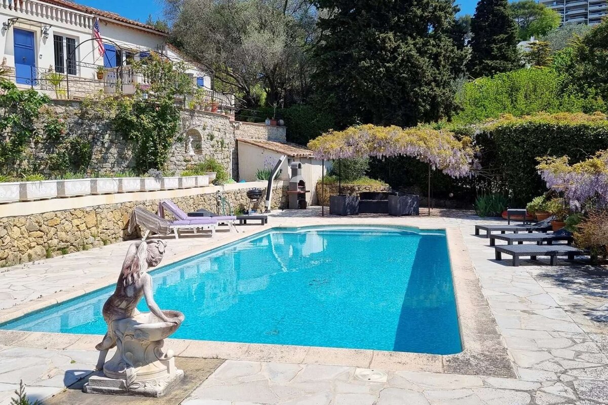 A sunny Mediterranean villa features a sparkling blue pool, terraced gardens, flowering wisteria providing shade, and inviting outdoor lounge areas.