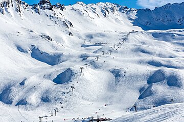 Vast snowy mountain ski resort with numerous ski lifts, groomed slopes, and skiers under a clear blue sky.