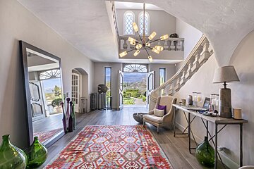 An elegant entryway with a vibrant geometric rug, a grand staircase, a modern chandelier, and open doors showing a scenic garden.