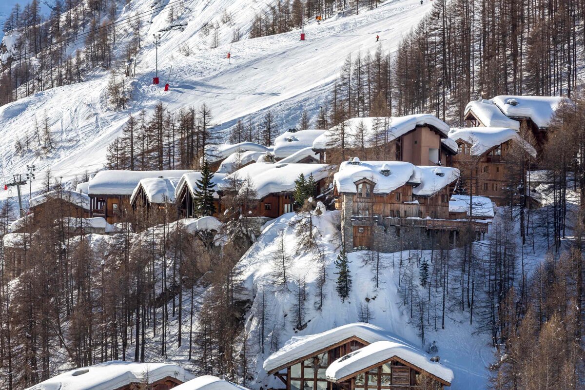 Charming snow-covered chalets on a steep, forested mountain, with a ski slope and lifts above.