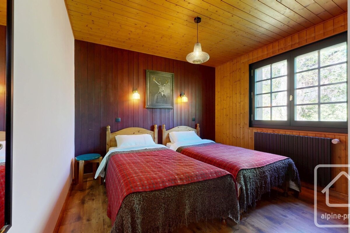 A cozy, rustic bedroom with two single beds, wood paneling on the walls and ceiling, a large window, and a framed deer portrait.