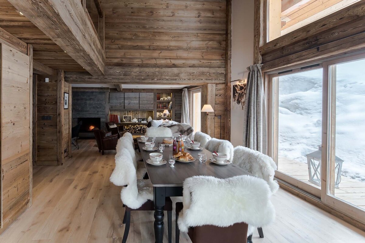 A cozy, rustic wooden chalet dining room with a set table, fluffy white chairs, and a warm fireplace, overlooking a snowy mountain landscape.