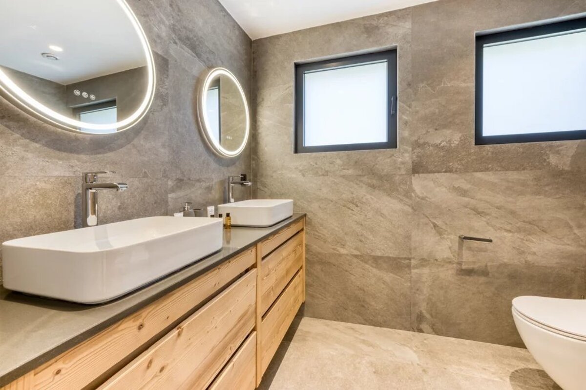 Modern bathroom with grey tile walls, a wooden double vanity with vessel sinks, glowing round mirrors, and frosted windows.