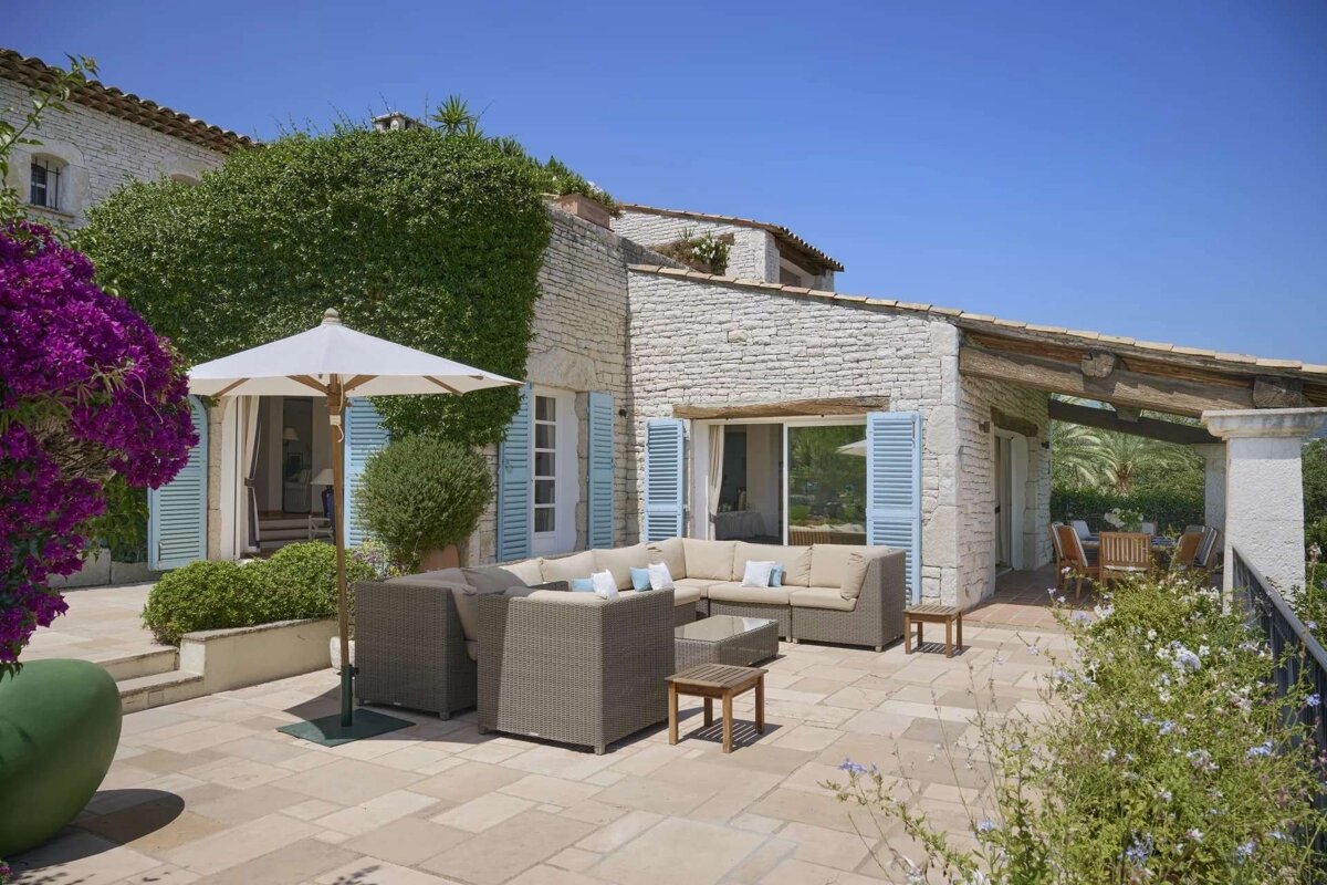 Sunny stone villa patio with a large wicker sectional sofa, white umbrella, blue shutters, and vibrant purple flowers under a clear sky.