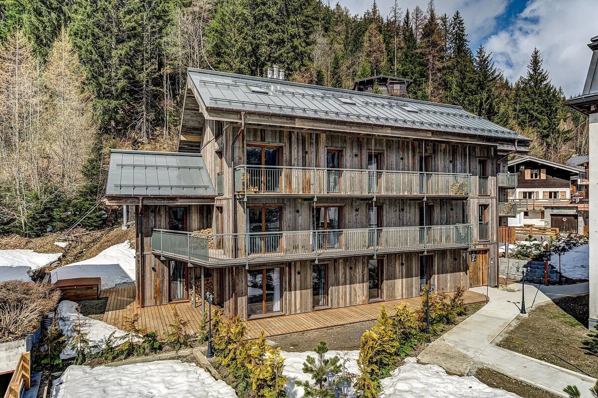 A large wooden chalet with multiple balconies and a metal roof stands on a snow-dusted hillside, backed by a dense forest under a cloudy sky.