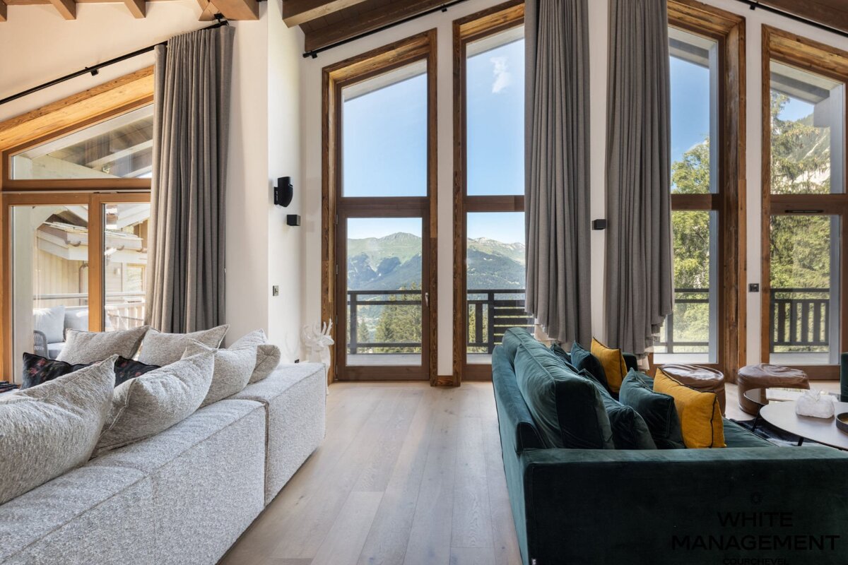 A modern living room with large, floor-to-ceiling windows offering stunning mountain views, featuring comfortable grey and green sofas.