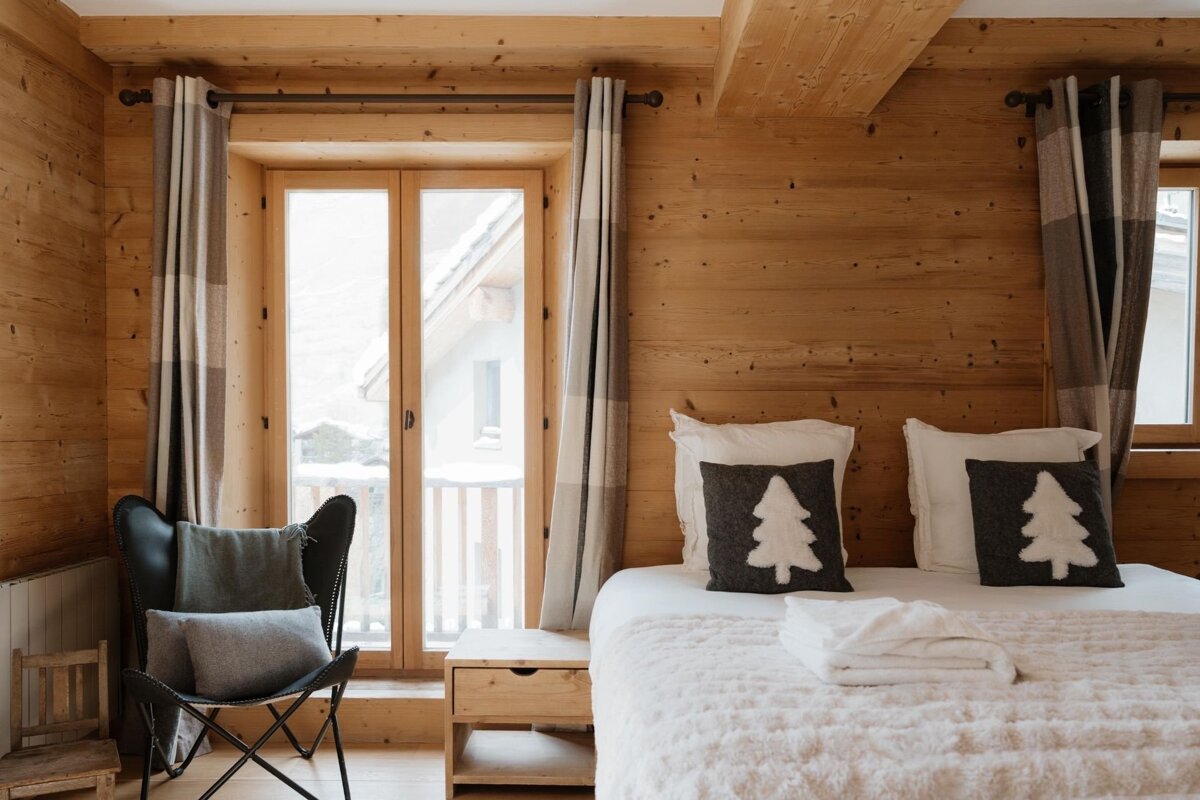 Cozy wooden cabin bedroom with pine tree pillows on the bed, a butterfly chair, and glass doors looking out to a snowy landscape.