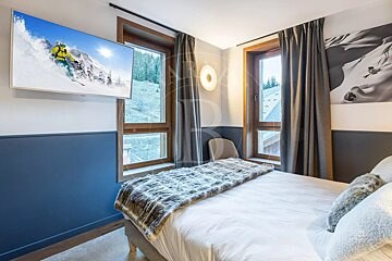 Modern alpine bedroom with a bed featuring a fur throw, two windows, blue-paneled walls, and a TV showing a skier.