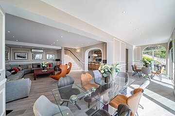 Spacious, modern open-plan living/dining room with a glass table, curved sofa, elegant staircase, and a bar in the background. Neutral decor.