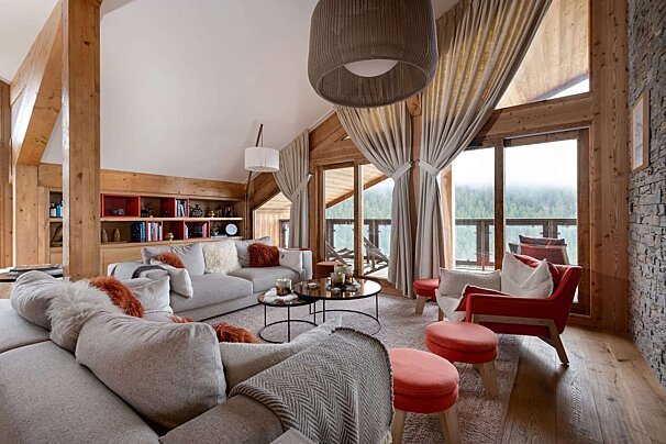 A cozy, wood-paneled chalet living room features a large grey sofa, red accents, and expansive windows with a forest view, creating a warm ambiance.
