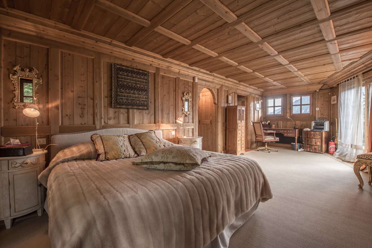 Cozy, wood-paneled chalet bedroom with a large plush bed, rustic wooden walls and ceiling, and a study area in the background.