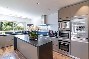 Bright, modern kitchen with an island, white orchids, stainless steel appliances (oven, wine fridge), and a large window overlooking greenery.