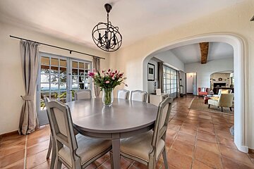 A charming dining room with a round table, fresh flowers, and French doors offering a scenic view, connected to a cozy living space.