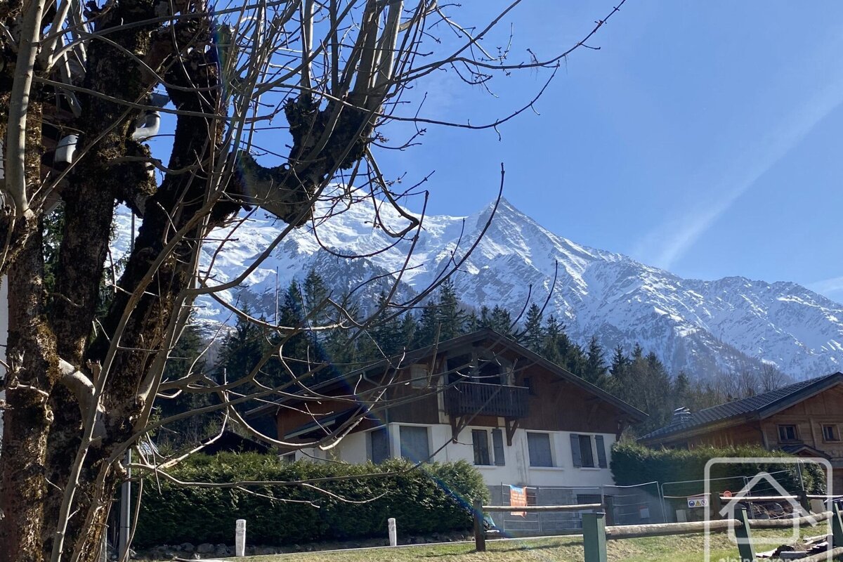 A snow-capped mountain rises behind a chalet-style house nestled among evergreen trees, with bare branches framing the foreground under a clear blue sky.