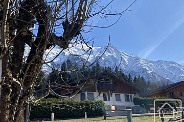 A snow-capped mountain rises behind a chalet-style house nestled among evergreen trees, with bare branches framing the foreground under a clear blue sky.