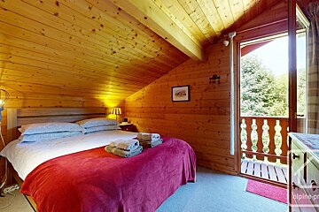 Cozy, wood-paneled attic bedroom with a bed, red blanket, and open door to a balcony overlooking lush green trees.