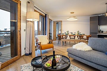 Cozy, modern chalet living room with a grey sofa, an orange armchair, and a dining table set in the background.