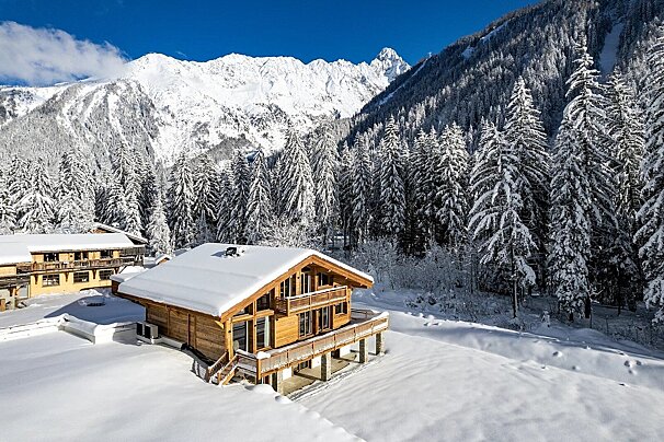 Wooden chalets covered in snow, nestled in a snowy pine forest with majestic snow-capped mountains under a bright blue sky.
