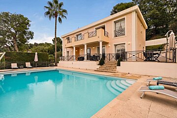 A large two-story peach house with balconies overlooks a swimming pool, with a tall palm tree and lush greenery under a clear sky.