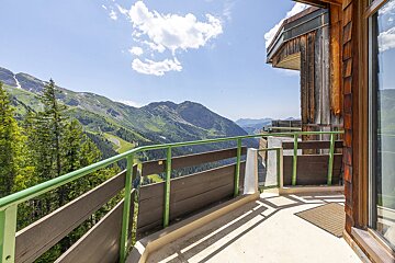 A wooden chalet balcony with a green railing overlooks a stunning view of lush green mountains under a blue sky.