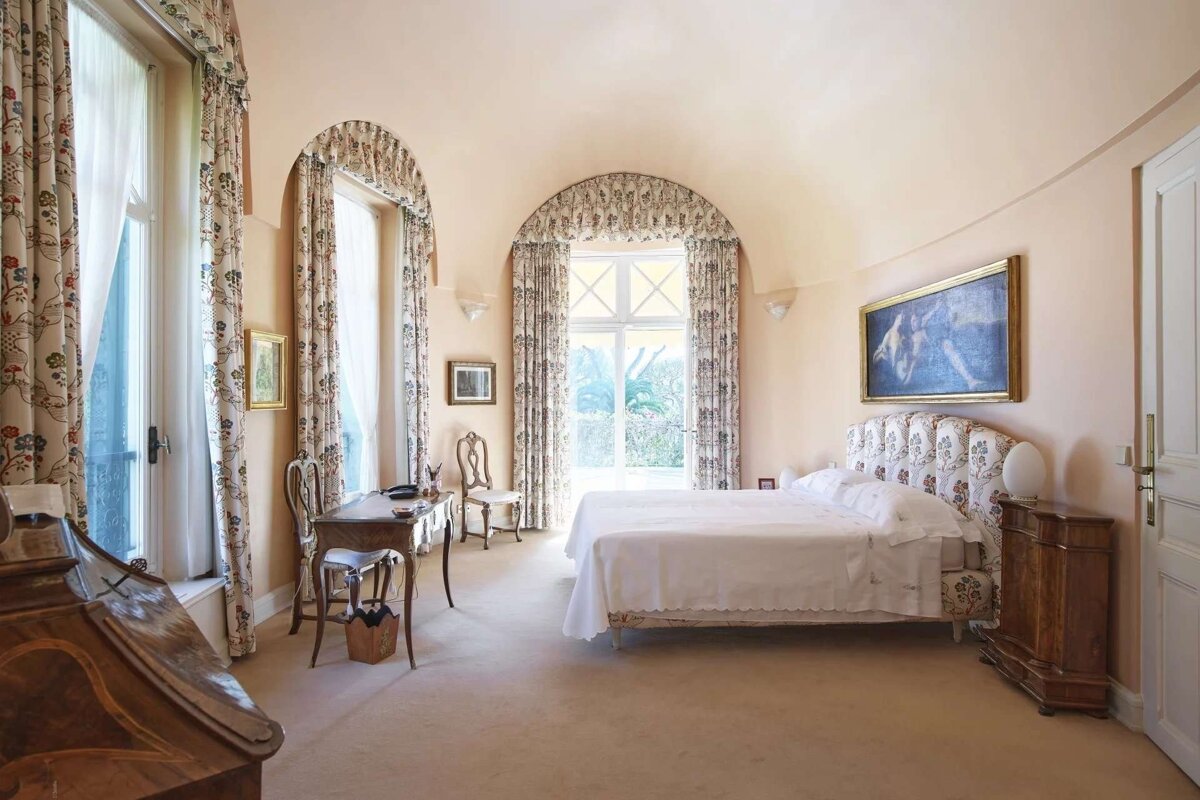 A spacious, elegant bedroom with pale peach walls, a vaulted ceiling, large arched windows with floral curtains, a bed, and antique furniture.