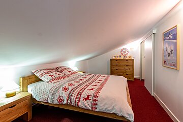 A cozy attic bedroom with a sloped ceiling, featuring a bed with red and white reindeer-patterned bedding, wooden furniture, and a red carpet.