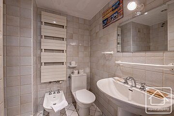 A light-colored tiled bathroom with a toilet, bidet, pedestal sink, towel warmer, mirror, and some wall art.