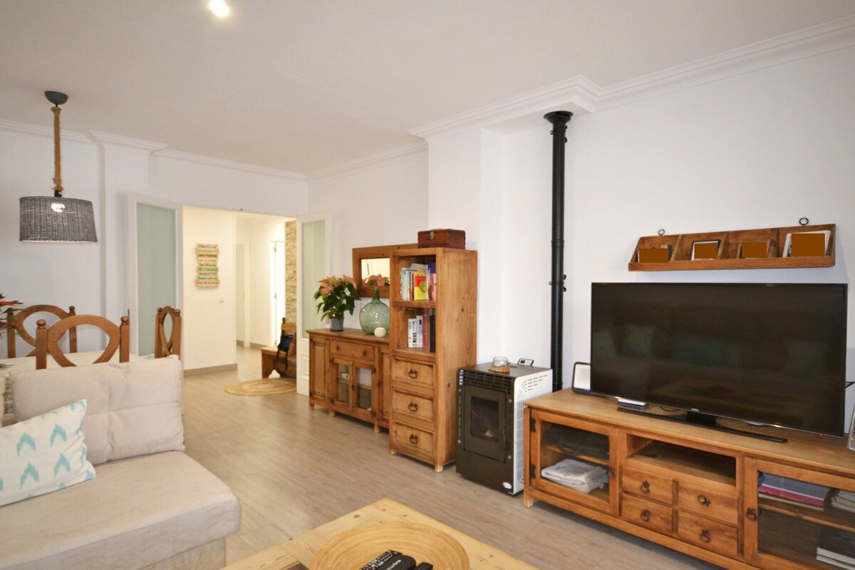 A bright living room with light wooden furniture, a TV, a couch, dining chairs, and a small stove, all on a light wooden floor.