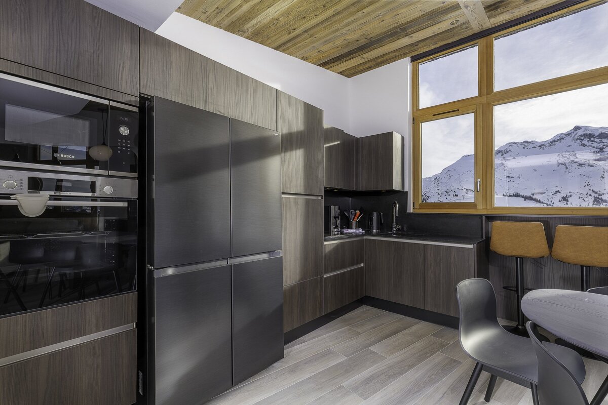 Modern dark kitchen with wood-grain cabinets and stainless appliances. Features a large window showcasing snow-covered mountains and a rustic wood plank ceiling.