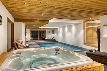 A sleek indoor swimming pool and glowing hot tub are set under a warm wood ceiling, creating a luxurious, modern spa retreat.