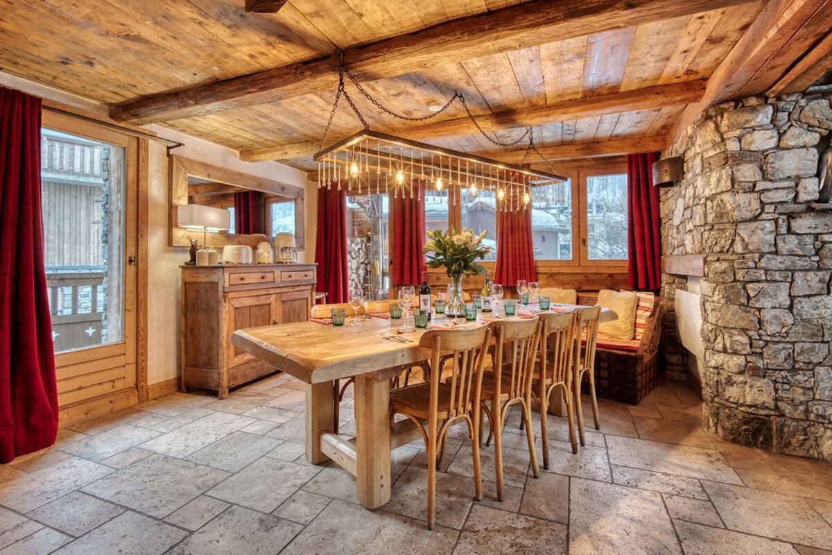 A rustic dining room with a long wooden table set for a meal, exposed wood beams, a stone wall, red curtains, and large windows.