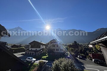 A bright sunburst shines over a village with houses and cars, framed by impressive mountains under a clear blue sky.