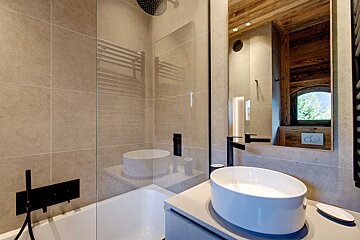 Sleek, modern bathroom with a bathtub/shower, vessel sink, and mirror reflecting a scenic window. Neutral tiles and black fixtures.
