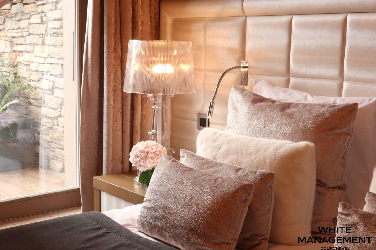 A luxurious bedroom features a bed with plush pillows, a glowing lamp, and pink flowers on a nightstand, beside a window revealing a stone wall outside.
