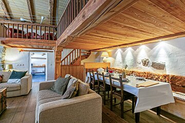Cozy rustic chalet interior featuring a wooden loft, a dining table set for dinner, comfortable sofas, and warm lighting.