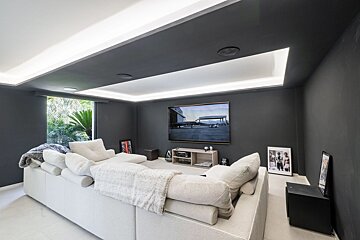 Modern media room with a large white sectional sofa, big TV on dark walls, and illuminated cove ceiling. A window reveals lush greenery.